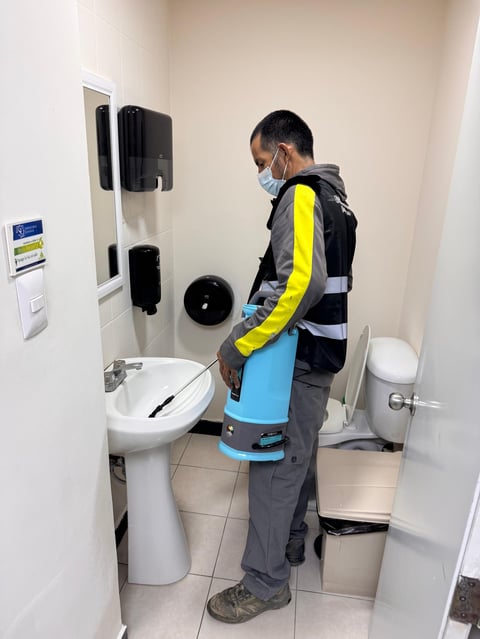Maintenance worker in yellow and black safety gear standing in a bathroom, holding cleaning equipment near a sink and toilet