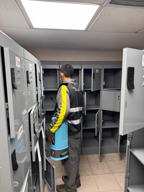 Worker in yellow safety vest standing in locker room with open metal lockers and fluorescent ceiling lights