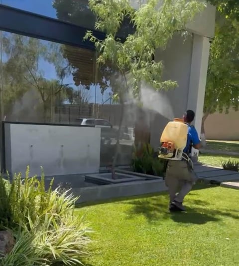 Person in blue shirt spraying water on modern house exterior with green lawn and landscaping