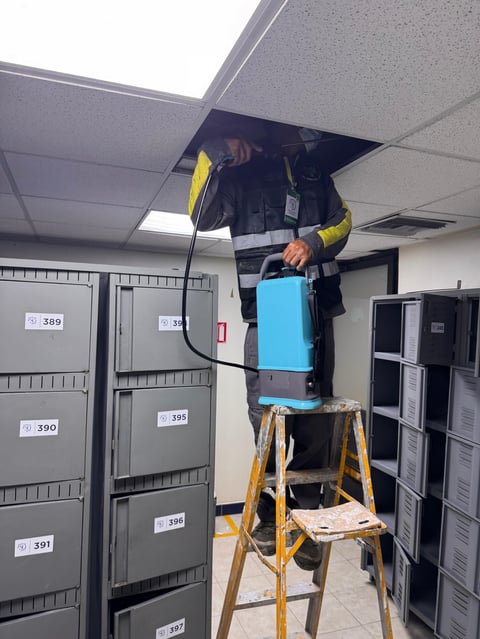 Electrician in yellow safety gear working on ceiling installation with blue equipment, standing on yellow ladder in technical room with electrical panels